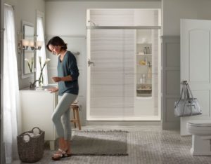 A woman stands clothed by the sink of her bathroom, which features a walk-in shower in the back